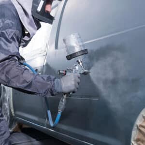 Peintre automobile en tenue de protection appliquant une couche de peinture grise sur une portière de voiture à l'aide d'un pistolet.
