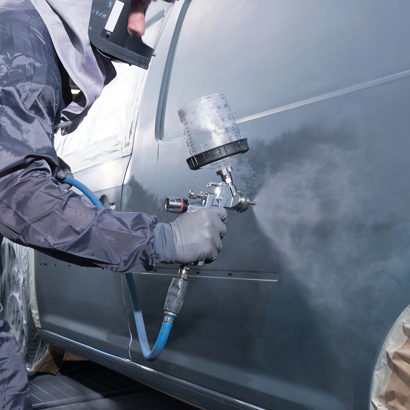 Peintre automobile en tenue de protection appliquant une couche de peinture grise sur une portière de voiture à l'aide d'un pistolet.