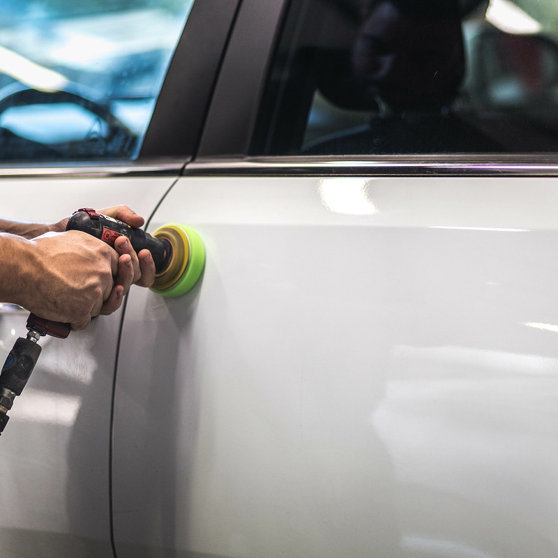 Mains polissant la porte blanche d'une voiture avec une polisseuse rotative verte, retirant les rayures et ravivant l'éclat.