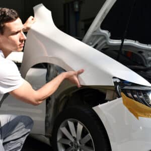 Carrossier installant une aile avant blanche sur une voiture en réparation. L'homme est concentré sur l'ajustement de la pièce.
