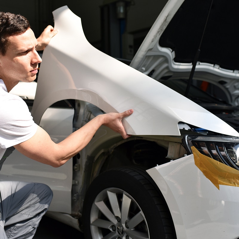 Carrossier installant une aile avant blanche sur une voiture en réparation. L'homme est concentré sur l'ajustement de la pièce.