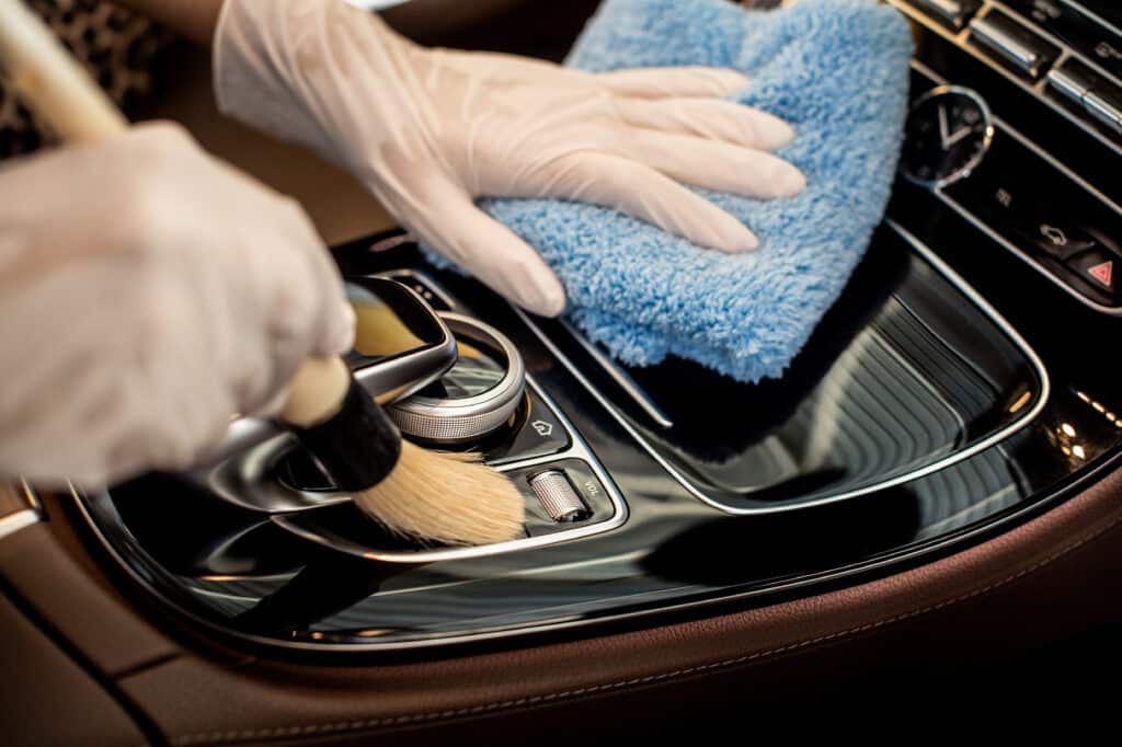 Mains gantées nettoyant méticuleusement l'intérieur d'une voiture de luxe avec une brosse et une microfibre, détaillant le tableau de bord et les commandes.