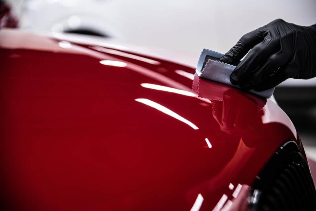Main gantée appliquant un produit de protection sur la carrosserie rouge brillante d'une voiture, pour un detailing impeccable.