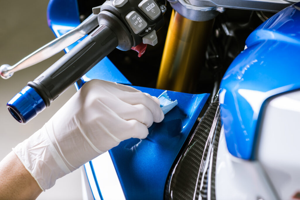Main gantée de blanc appliquant avec un tampon un traitement sur le carénage bleu métallisé d'une moto, près du guidon et carbone.