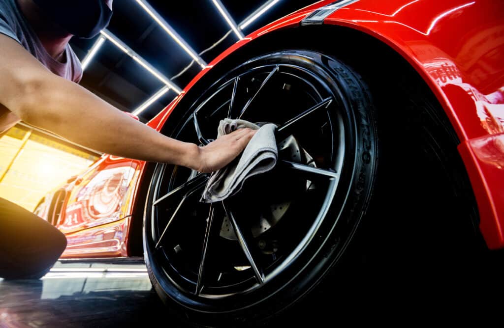 Homme masqué nettoyant la jante noire et brillante d'une voiture de sport rouge avec un chiffon. Détail auto professionnel, pneu Pirelli.