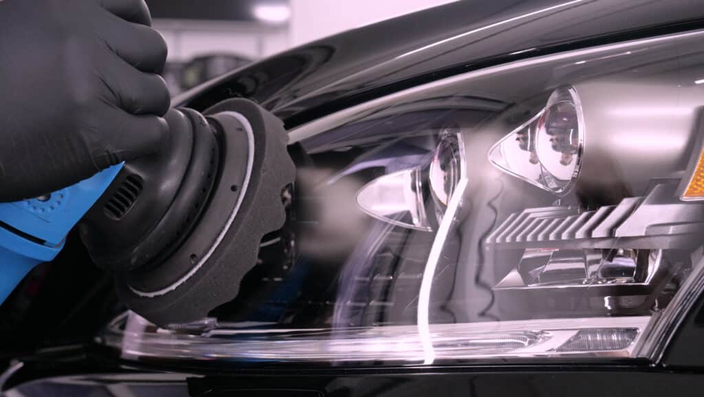 Main gantée polissant le phare d'une voiture noire avec une machine bleue et un pad en mousse. La carrosserie est brillante.