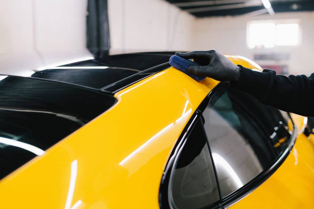 Main gantée appliquant un revêtement céramique sur le toit jaune vif et brillant d'une voiture, dans un atelier de detailing.