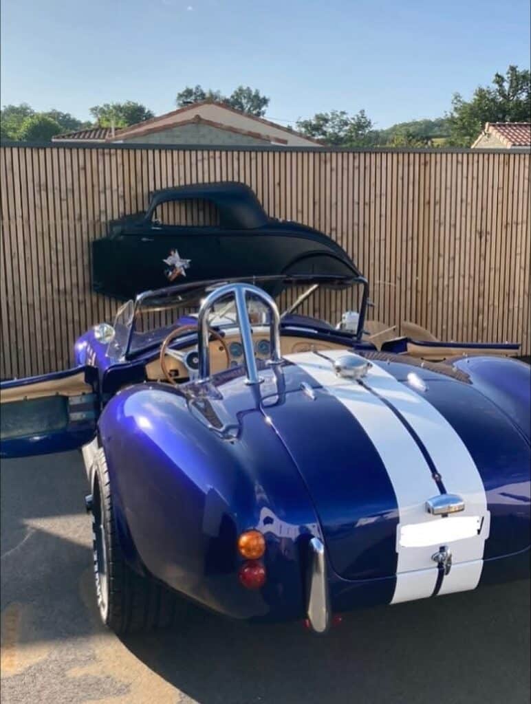 AC Cobra bleu éclatant avec bandes blanches, portes ouvertes, devant une palissade en bois où est accrochée une carrosserie noire.
