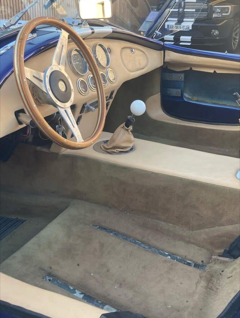 Intérieur d'un roadster bleu style Cobra avec tableau de bord beige, volant bois, levier de vitesse à boule blanche et moquette marron. Plaque AR-163-CJ visible.